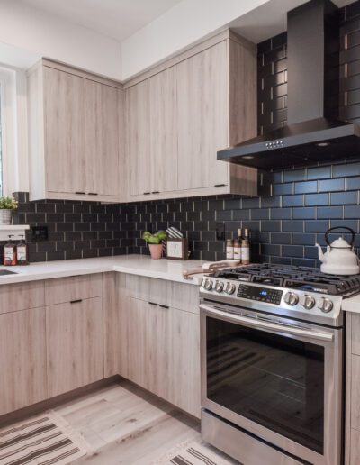 Modern kitchen with light wood cabinets, black subway tile backsplash, stainless steel oven, and a window above the sink. A kettle sits on the stove, and striped rugs cover the floor.