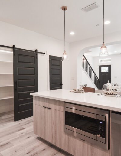 Modern kitchen with light wood flooring, a large island featuring a microwave, hanging pendant lights, and sliding pantry doors. A staircase and seating area are visible in the background.