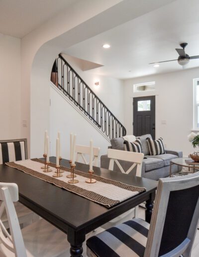 Modern living and dining area with black furniture, striped chairs, a gray sofa, and a staircase. White walls, large windows, and a ceiling fan enhance the space. Candlesticks decorate the table.