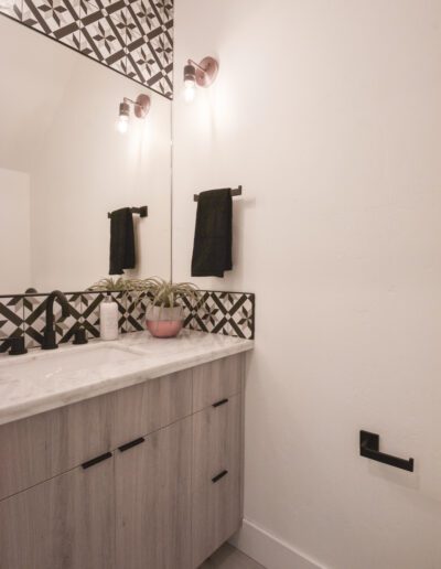 Modern bathroom with a wooden vanity, marble countertop, geometric mirror frame, pink potted plant, and black fixtures.