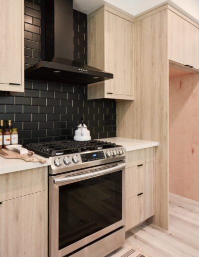 Modern kitchen with light wood cabinets, black tile backsplash, stainless steel oven, and a black range hood. A barn door leads to an adjacent room. Light wood flooring throughout.