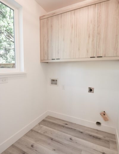 Empty laundry room with light wood cabinets, gray flooring, and utility hookups below. A window on the left side shows greenery outside.