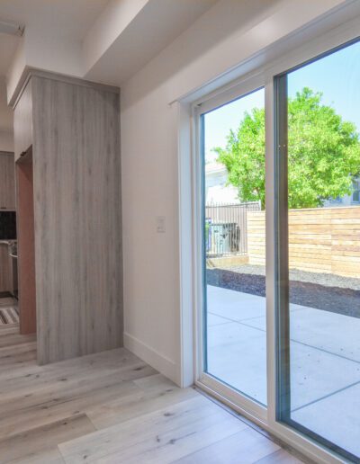 Bright room with wood flooring, sliding glass door leading to a patio, and a view of a fenced yard with a tree. Interior includes a modern kitchen and seating area.