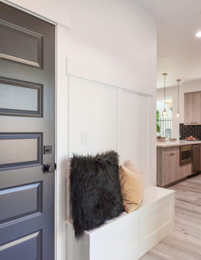 Interior of a home with a black door, a bench with cushions, and a view into a modern kitchen with light wood cabinets and hanging lights.