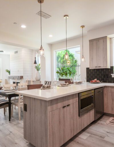 Modern open-plan kitchen and living area with wooden cabinets, pendant lights, a dining table, and a sofa; large windows provide natural light.