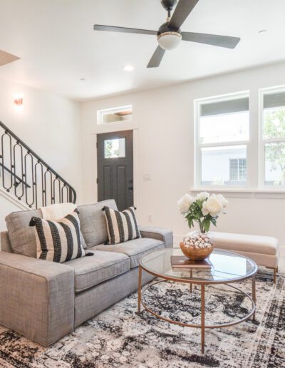 Modern living room with a gray sofa, decorative pillows, a round glass coffee table, and a patterned rug. Features a staircase with black railing, ceiling fan, and fireplace.