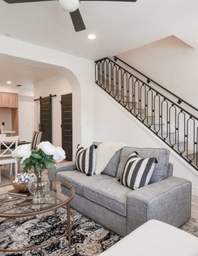 Modern open-concept living room with a gray sofa, patterned rug, and flowers. In the background, a kitchen with wooden cabinets and a black accent wall. Staircase with black railing on the right.