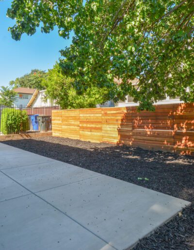 A fenced backyard with a concrete patio, wooden privacy screen, and surrounding trees under a clear blue sky.