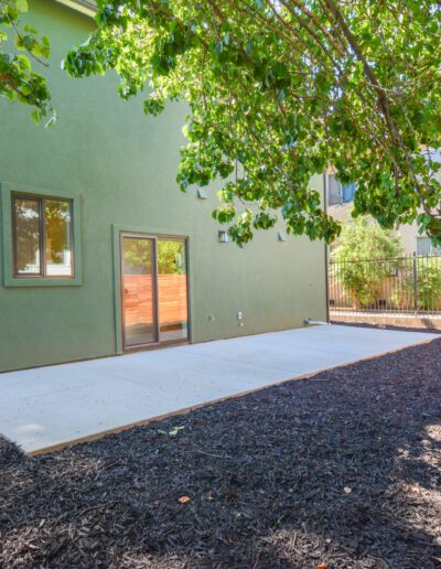 A green building with a sliding glass door opens to a concrete patio, surrounded by a mulched yard and trees.