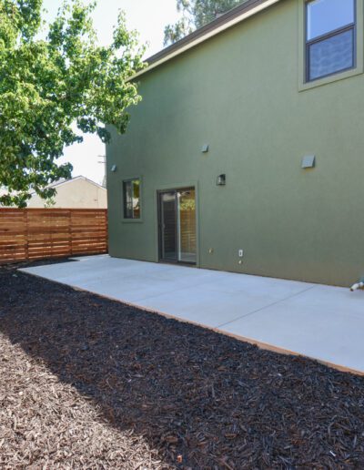 A two-story green house with a concrete patio, wooden fence, and mulched ground area. A large tree stands near the corner of the house.