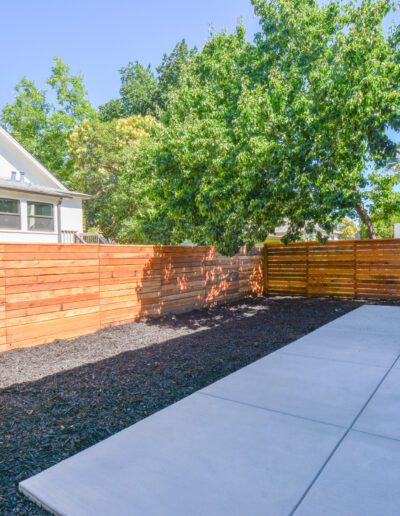 A backyard with a wooden fence, dark mulch area, and a concrete patio. There are trees surrounding the fence.