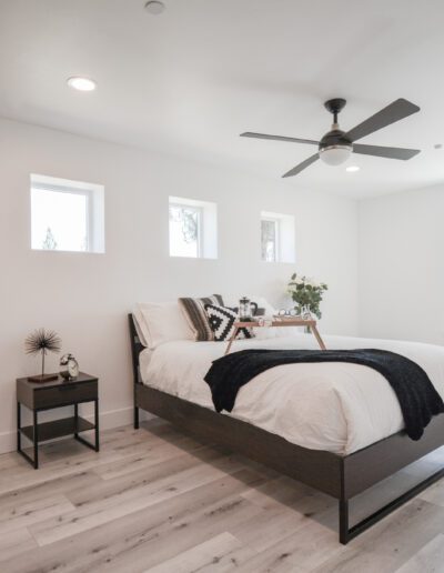 Minimalist bedroom with a bed, white bedding, black accents, a side table, and a ceiling fan. natural light enters through three small windows and one large window. Wood flooring throughout.