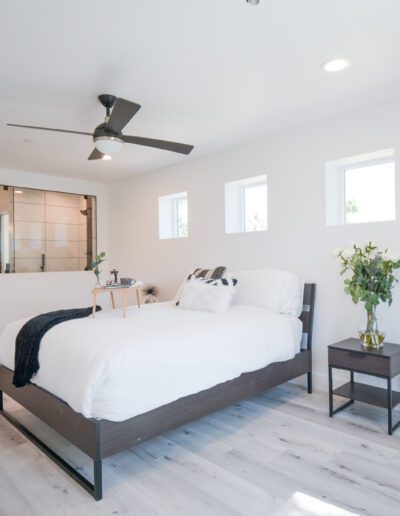 Modern bedroom with a large bed, ceiling fan, and three small windows. Hardwood flooring, a bedside table with flowers, and an open bathroom with a shower in the background.