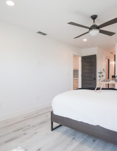 Bright bedroom with a large bed, a ceiling fan, and light wood flooring. A tray with breakfast items is placed on the bed. A door leads to an adjoining room.