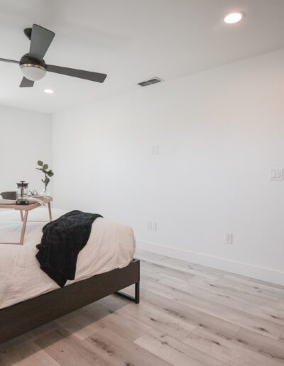 Minimalist bedroom with a dark wood bed, white bedding, and a tray on top. A ceiling fan is above, and light wood flooring extends throughout. A doorway leads to another room.