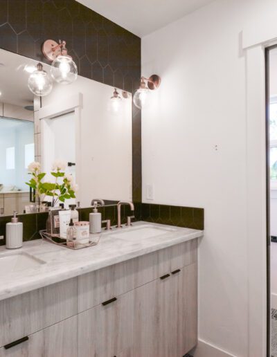 Modern bathroom with two sinks and a large mirror above. Light fixtures are mounted on a dark hexagonal tiled wall. The room features white walls and a small window.