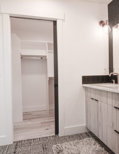 Modern bathroom with light wood cabinetry, marble countertop, and a geometric black wall. A glass shower is on the left, and decorative jars and flowers are on the counter.