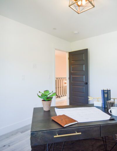 Modern home office with a dark desk, notebooks, a coffee cup, and potted plants. The door is ajar, revealing a hallway with a railing. The room is bright with light from a ceiling fixture.