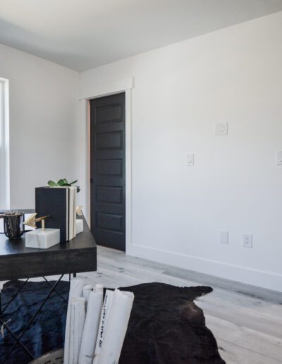 A minimalist home office with a black desk, a cow-print chair, and light wood flooring. A dark door and window are on one wall. Rolled-up papers and a plant are on the desk.