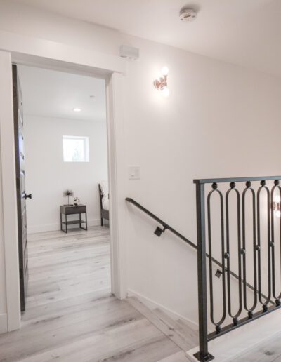 A bright hallway with light wood flooring, a black metal railing, and open doors leading to rooms.