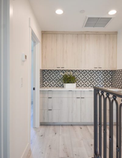 Modern hallway with light wood cabinets, patterned backsplash, and indoor plants. A black railing and adjacent bathroom with pendant lights are visible.