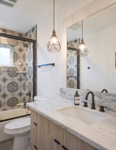 Modern bathroom with patterned tile, glass-enclosed bathtub, marble countertop, geometric pendant lights, and potted plants by the sink.