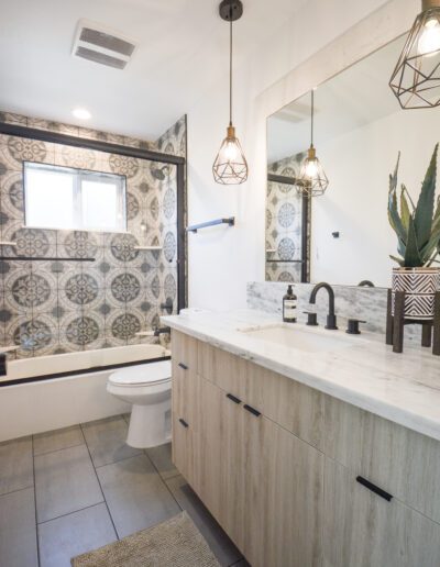 Modern bathroom with a tiled shower, wooden vanity, dual sinks, large mirror, geometric pendant lights, and a small plant.