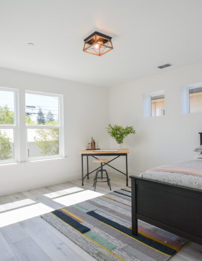 Bright, minimalist bedroom with a single bed, wooden desk, and colorful rug. Large windows let in natural light.