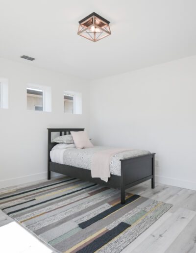 Minimalist bedroom with a single bed, desk, stool, and a patterned rug. Wall features small windows. An adjacent doorway leads to a bathroom.