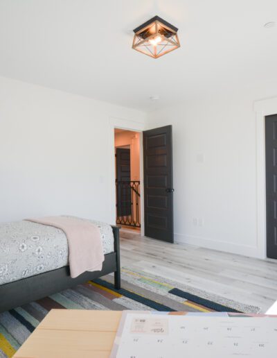 Minimalist bedroom with a single bed, patterned rug, and a wooden table. Dark sliding closet doors and a geometric ceiling light enhance the modern design.