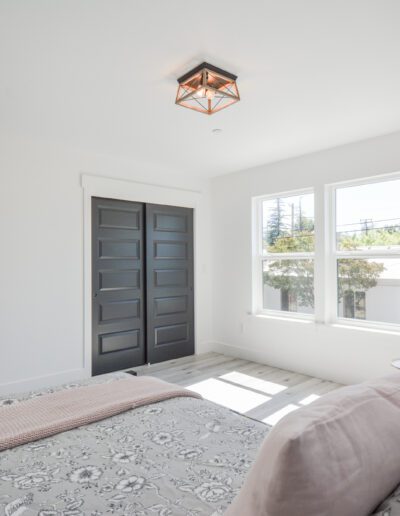 Bright bedroom with a patterned bedspread, large windows, a small desk with a plant, and dark doors.