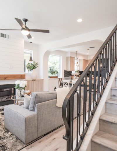 Modern living room with light wood floors, a gray sofa, and a small fireplace. Staircase with a black railing on the right, leading to a second floor. Dining area visible in the background.