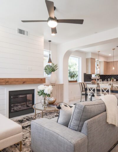 Modern living room with a gray sofa, fireplace, and open kitchen. Light wood floors, ceiling fan, and pendant lights. Simple decor with plants and neutral colors.