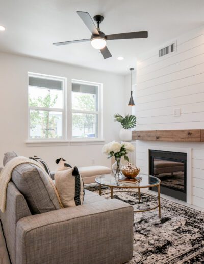 A modern living room with a gray sofa, patterned rug, and a white shiplap accent wall with a fireplace. Large windows, ceiling fan, and a dark front door are also visible.