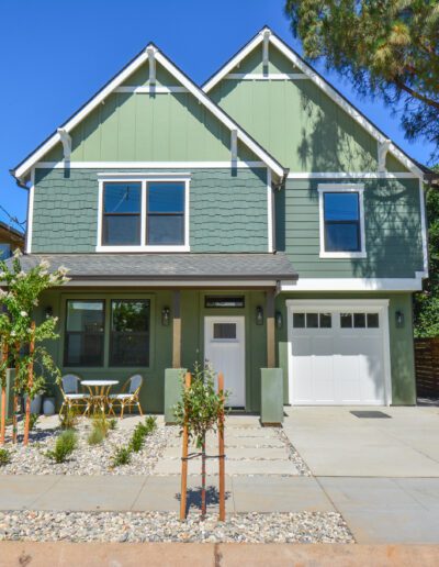 A two-story green house with a small front yard, adjacent trees, and a driveway.