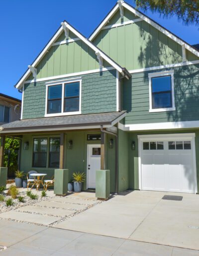 A two-story green house with white trim, featuring a garage, small front porch, and a landscaped front yard with a seating area and plants.