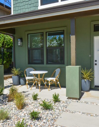 Front porch with a green exterior, white door, two chairs, and a small round table. Pebble-lined garden features plants and a tree.