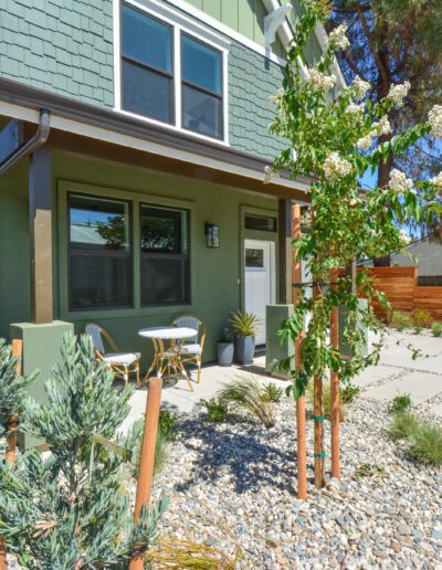 Modern house exterior with green siding, two-story windows, a small patio with chairs, and a drought-resistant garden featuring young trees and gravel landscaping.